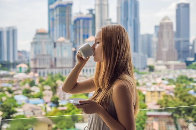 La giovane donna beve il caffè al mattino sul balcone con vista sulla grande città e sui grattacieli