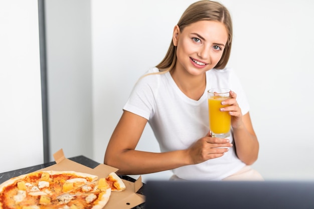 La giovane donna beve succo di frutta e mangia pizza e guarda il laptop in cucina