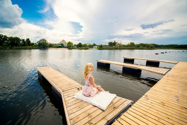 La giovane donna bionda affascinante si siede da solo su un molo di legno e guarda il fiume.