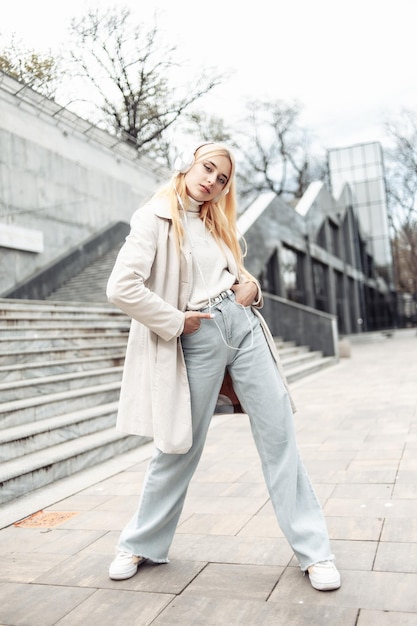 La giovane donna bionda alla moda in trench ascolta musica sulle cuffie all'aperto