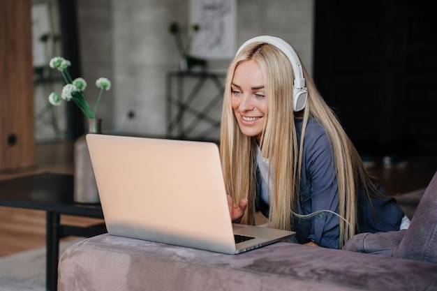 La giovane donna bionda allegra nei sorrisi delle cuffie si siede sul divano con il computer portatile fa sorrisi di videochiamata