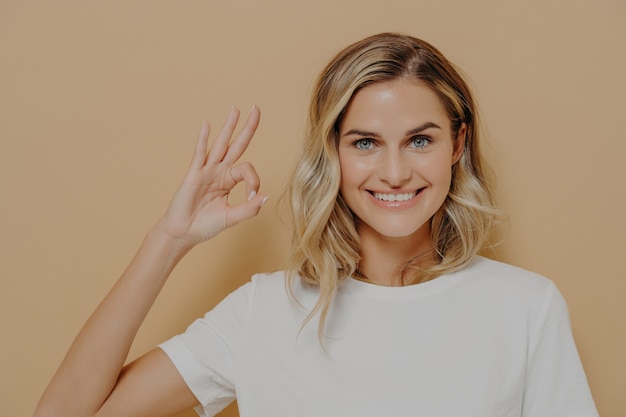 La giovane donna bionda carina positiva in maglietta bianca mostra il segno OK con una mano, sorridendo felicemente alla telecamera, la femmina che mostra tutto è super e bene. Linguaggio del corpo ed emozioni umane