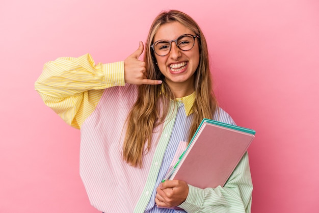 La giovane donna bionda caucasica dello studente tiene i libri isolati mostrando un gesto di chiamata di telefono cellulare con le dita