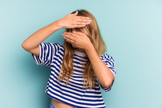 La giovane donna bionda caucasica isolata su fondo blu lampeggia alla macchina fotografica attraverso le dita, imbarazzata che copre il viso.