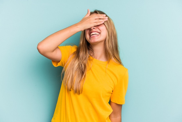 La giovane donna bionda caucasica isolata su fondo blu ride con gioia tenendo le mani sulla testa. Concetto di felicità.