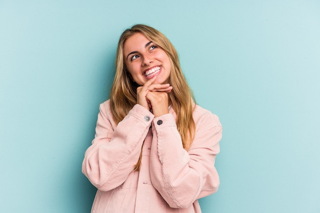 La giovane donna bionda caucasica isolata su fondo blu tiene le mani sotto il mento, sta guardando felicemente da parte.