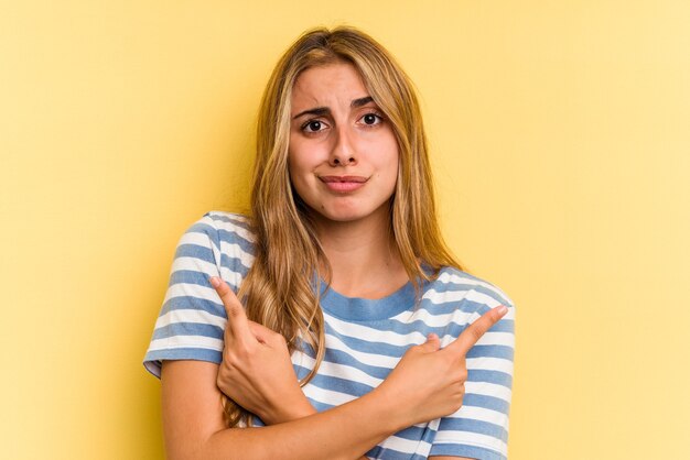 La giovane donna bionda caucasica isolata su sfondo giallo punta lateralmente, sta cercando di scegliere tra due opzioni.