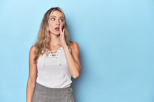 La giovane donna bionda caucasica nello studio blu sta dicendo una notizia segreta calda frenante e guardando da parte