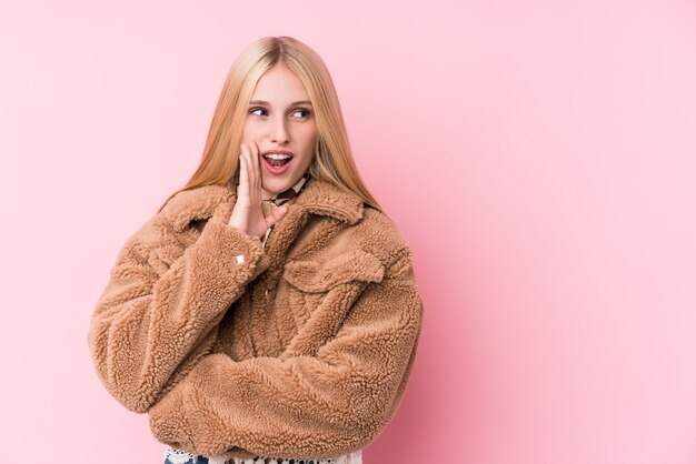 La giovane donna bionda che indossa un cappotto sul rosa sta dicendo una notizia segreta di frenata calda e sta guardando da parte