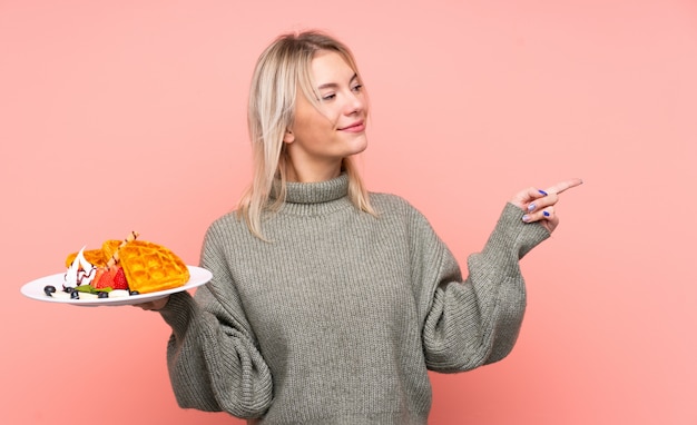 La giovane donna bionda che tiene le cialde sopra la parete rosa isolata che indica il lato per presentare un prodotto