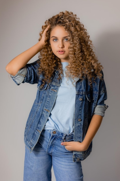 La giovane donna bionda dagli occhi azzurri con i capelli ricci in una giacca di jeans e jeans si erge su uno sfondo grigio