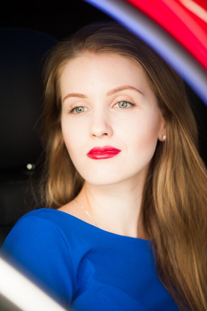 La giovane donna bionda europea di bello modo che si siede in macchina rossa si è vestita in vestito lungo blu