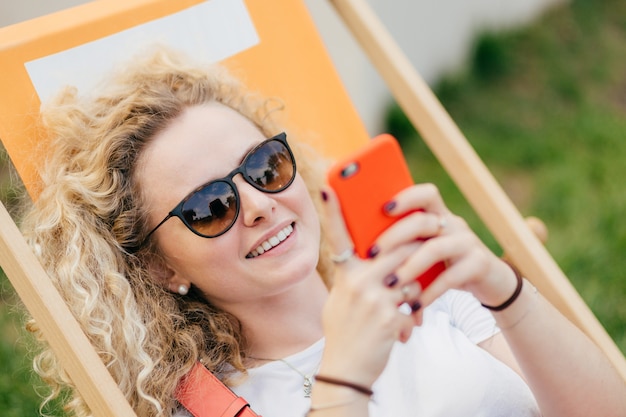 La giovane donna bionda riccia si trova in un'amaca, gode di una giornata estiva, blog e chat tramite smartphone