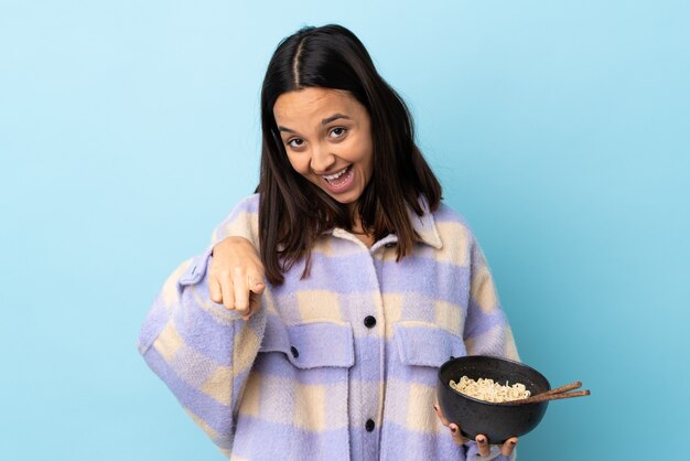 La giovane donna bruna di razza mista sopra il muro blu isolato punta il dito contro di te con un'espressione sicura mentre tiene una ciotola di spaghetti con le bacchette.