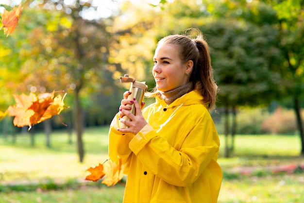 La giovane donna cammina nel parco in autunno dorato una ragazza in un impermeabile beve il tè da un comfort di thermos