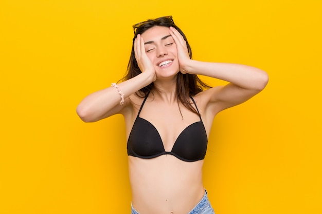 La giovane donna castana che porta un bikini su giallo ride con gioia tenendo le mani sulla testa.