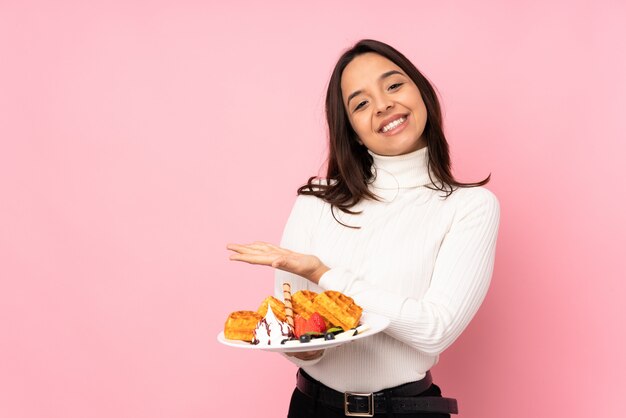 La giovane donna castana che tiene le cialde sopra la parete rosa isolata che presenta un'idea mentre sembra sorridere verso