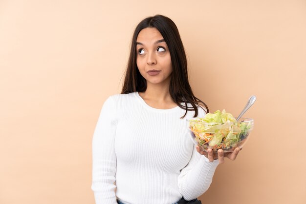 La giovane donna castana che tiene un'insalata che fa i dubbi gesticola lo sguardo laterale