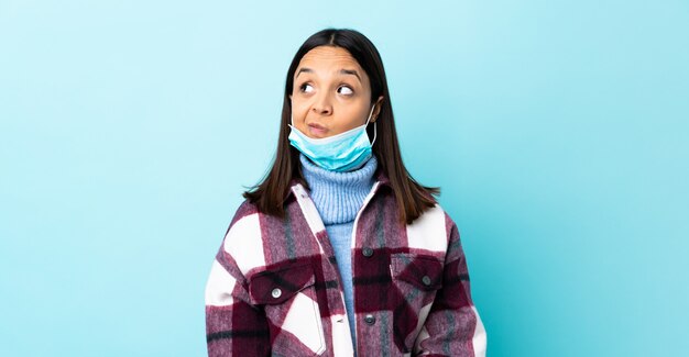 La giovane donna castana della corsa mista che protegge con una maschera e guanti sopra la parete blu che fa i dubbi gesturing lo sguardo laterale