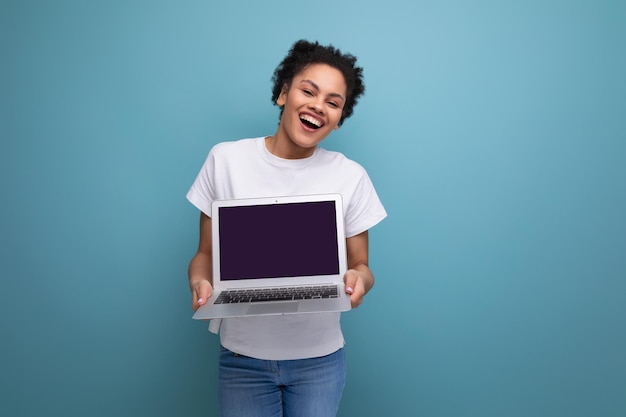 La giovane donna castana felice con i capelli afro in una canottiera bianca tiene in mano un computer portatile per lavorare