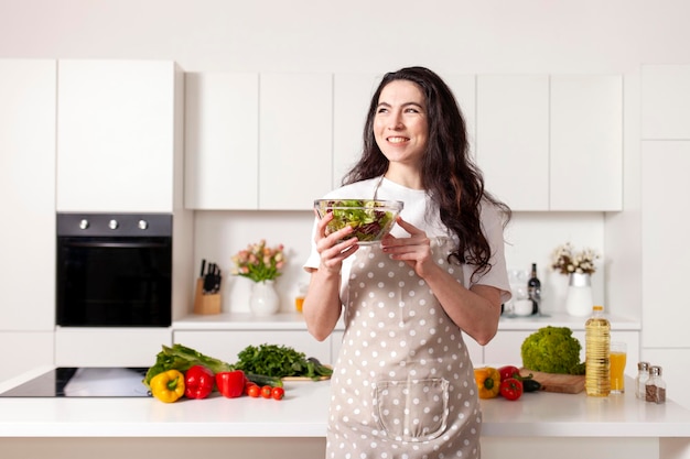 La giovane donna castana in grembiule tiene l'insalata vegetariana nella cucina moderna bianca e guarda di lato