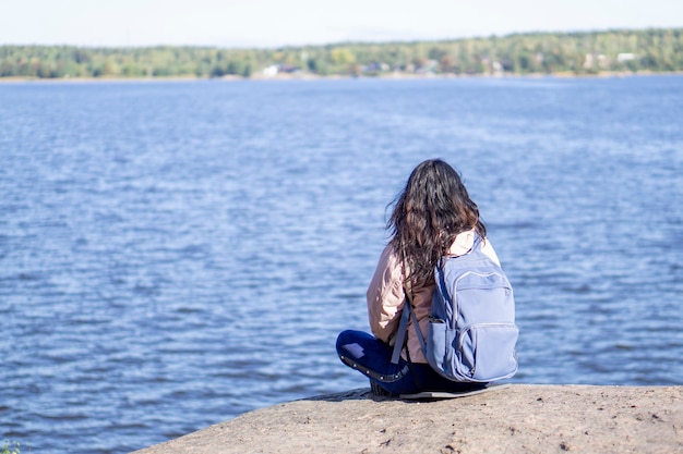 La giovane donna castana si siede con le spalle alla telecamera, lo zaino dietro le spalle e guarda il panorama del lago. Concetto di relax in natura