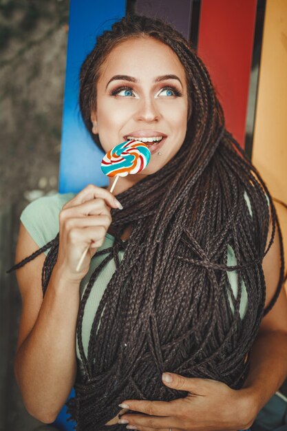 La giovane donna caucasica allegra con bei capelli lunghi intrecciati afro sta leccando il lecca-lecca, sorridendo e distogliendo lo sguardo.