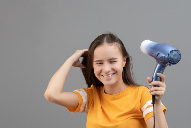 La giovane donna caucasica asciuga i capelli con un asciugacapelli su uno sfondo grigio divertendosi