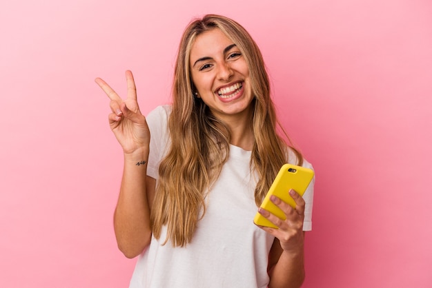 La giovane donna caucasica bionda che tiene un telefono cellulare giallo ha isolato gioiosa e spensierata che mostra un simbolo di pace con le dita.