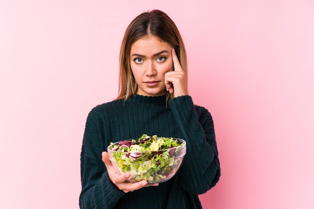 La giovane donna caucasica che giudica un'insalata isolata indicando il suo tempio con il dito, pensando, si è concentrata su un compito.