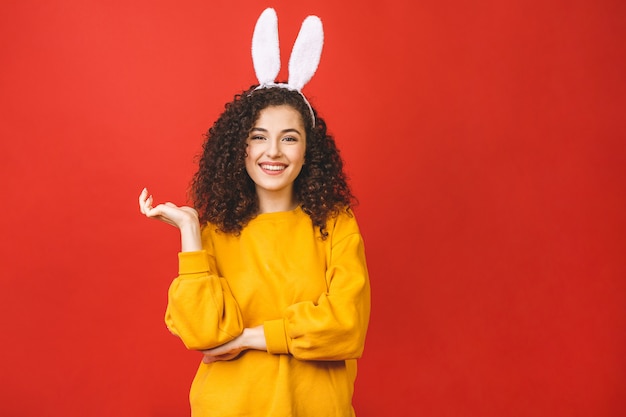 La giovane donna caucasica che indossa le orecchie di coniglio svegli di pasqua su rosso ha isolato il fondo mentre sorrideva sicuro e felice.
