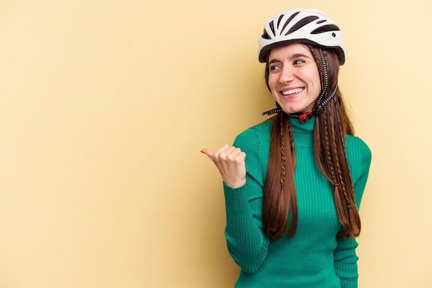 La giovane donna caucasica che indossa una bici da casco isolata su sfondo giallo indica con il pollice lontano, ridendo e spensierata.