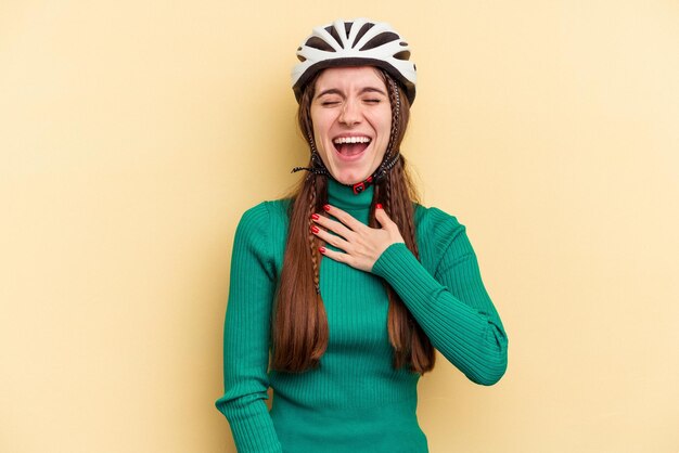 La giovane donna caucasica che indossa una bici da casco isolata su sfondo giallo ride ad alta voce tenendo la mano sul petto