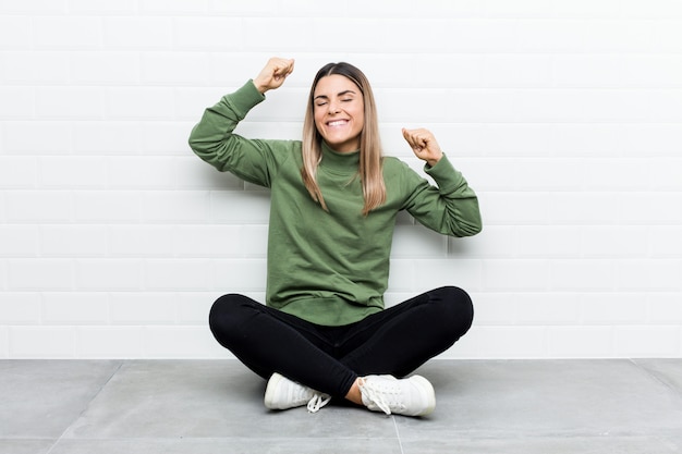 La giovane donna caucasica che si siede sul pavimento che celebra un giorno speciale, salta e alza le braccia con energia.