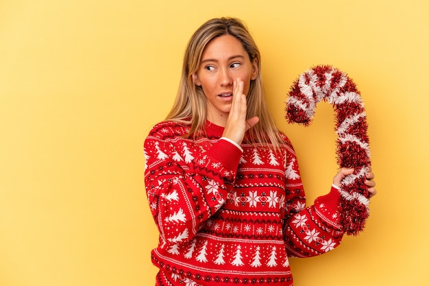 La giovane donna caucasica che tiene in mano un grosso bastone di natale isolato su sfondo giallo sta dicendo una notizia segreta di frenata calda e sta guardando da parte