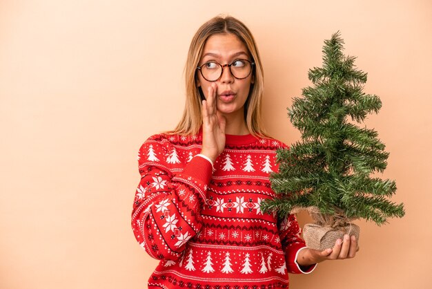 La giovane donna caucasica che tiene in mano un piccolo albero di natale isolato su sfondo beige sta dicendo una notizia segreta di frenata calda e sta guardando da parte