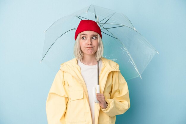La giovane donna caucasica che tiene l'ombrello isolato su sfondo blu confusa si sente dubbiosa e insicura