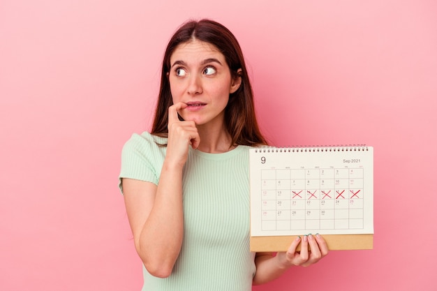 La giovane donna caucasica che tiene un calendario isolato su sfondo rosa si è rilassata pensando a qualcosa guardando uno spazio di copia.