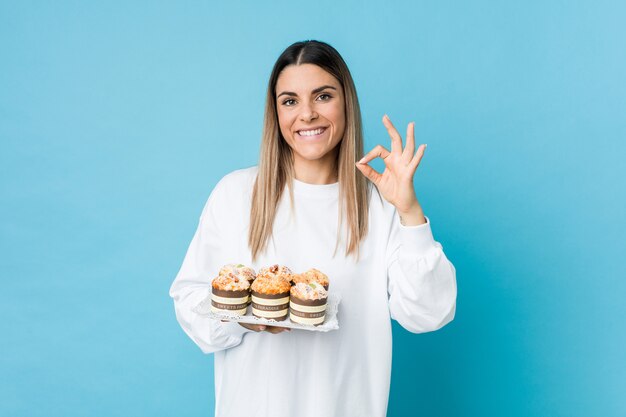 La giovane donna caucasica che tiene un dolce agglutina il gesto giusto di mostra allegro e sicuro.