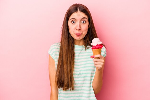 La giovane donna caucasica che tiene un gelato isolato su sfondo rosa alza le spalle e apre gli occhi confusi.