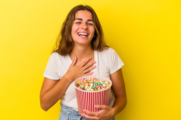 La giovane donna caucasica che tiene un popcorn isolato su sfondo giallo ride ad alta voce tenendo la mano sul petto.