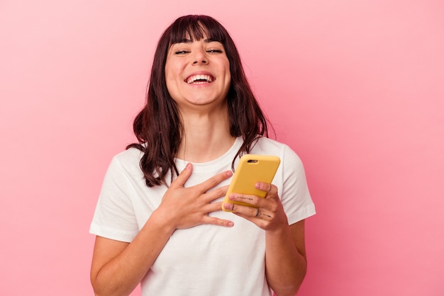 La giovane donna caucasica che tiene un telefono cellulare isolato su sfondo rosa ride ad alta voce tenendo la mano sul petto.