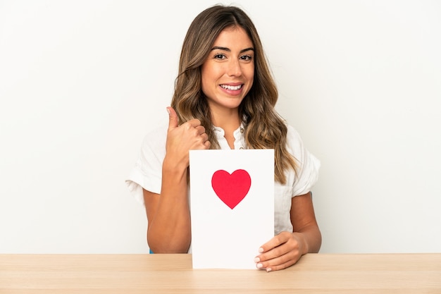 La giovane donna caucasica che tiene una carta di San Valentino ha isolato sorridendo e alzando il pollice in su