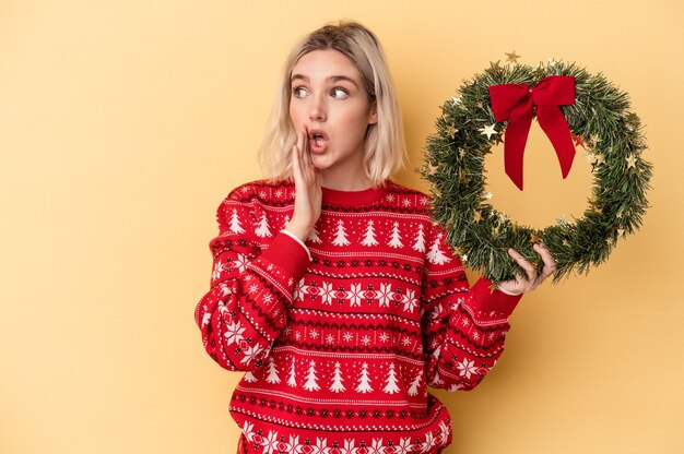 La giovane donna caucasica che tiene una ghirlanda di Natale isolata su sfondo giallo sta dicendo una notizia segreta di frenata calda e sta guardando da parte