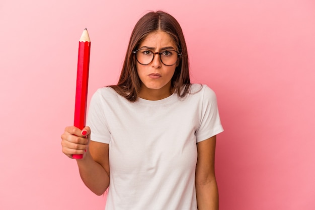 La giovane donna caucasica che tiene una grande matita isolata su fondo rosa alza le spalle e apre gli occhi confusi.