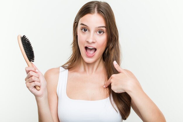 La giovane donna caucasica che tiene una spazzola per capelli ha sorpreso indicando se stesso, sorridendo ampiamente.