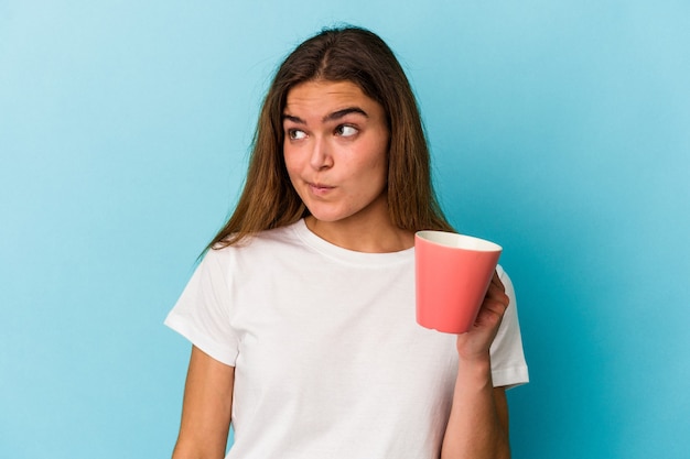 La giovane donna caucasica che tiene una tazza isolata su sfondo blu confusa, si sente dubbiosa e insicura.