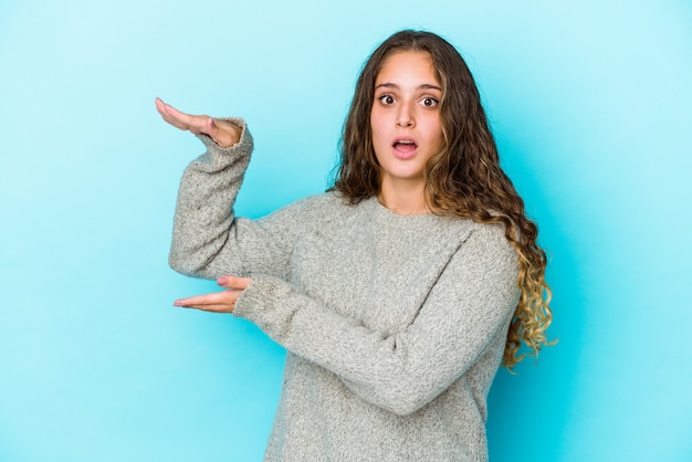 La giovane donna caucasica dei capelli ricci ha isolata scioccata e stupita tenendo uno spazio della copia tra le mani.