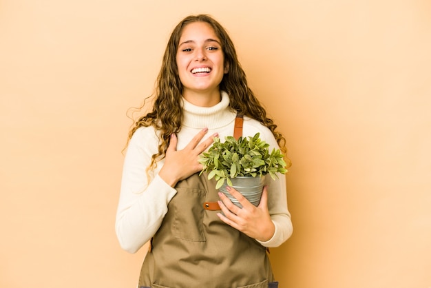 La giovane donna caucasica del giardiniere che tiene una pianta isolata ride ad alta voce mantenendo la mano sul petto.