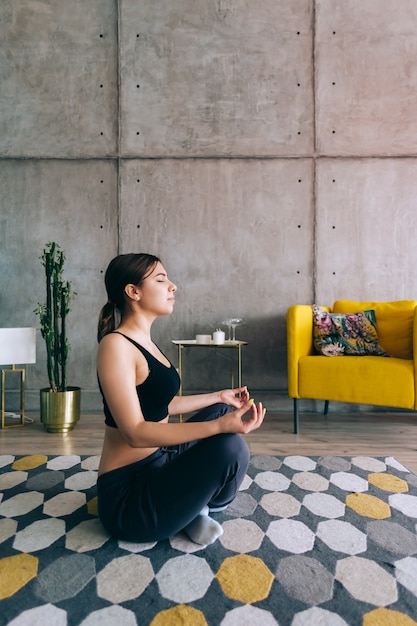 La giovane donna caucasica di forma fisica medita, facendo yoga al chiuso a casa vicino al letto. Rimanere in forma e in salute